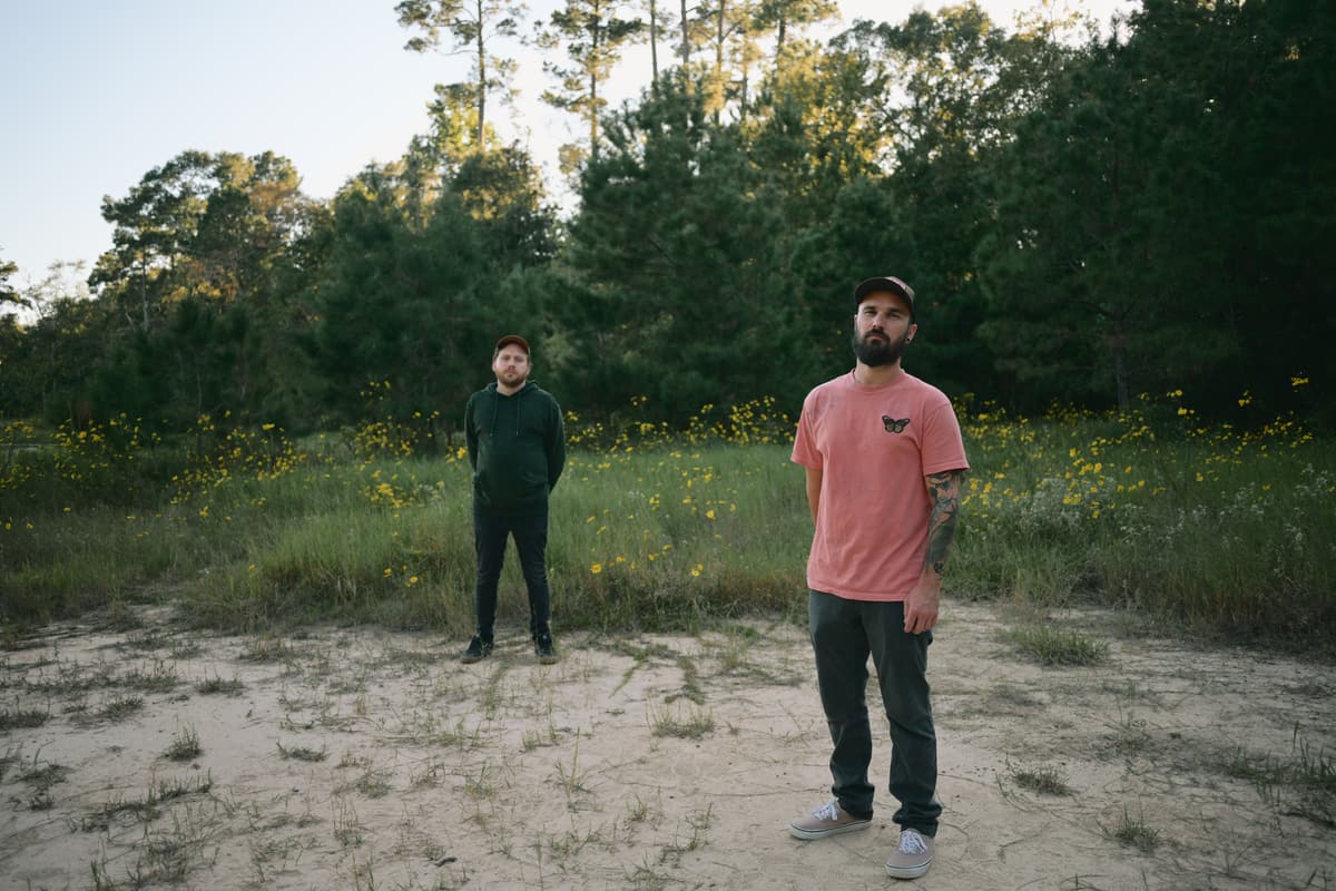 two of the band standing apart in a sandy clearing in front of pine trees